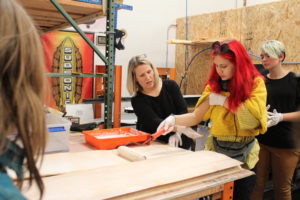 A student painting a piece of wood with guidance from a business owner.