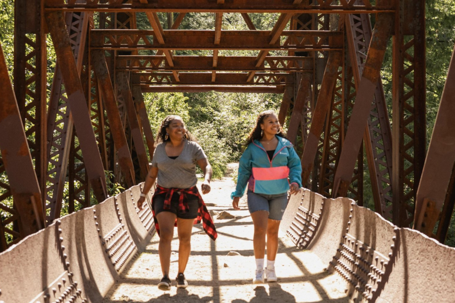 Two women walk side by side toward the camera through a rustic steel truss bridge, smiling and chatting. One wears a gray t-shirt with a flannel tied around her waist; the other wears a teal and pink pullover and denim shorts. Lush green forest is visible through the far end of the bridge.