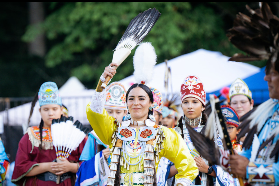 A woman in elaborate powwow regalia — including a yellow fringed dress, intricate beadwork breastplate, layered shell necklaces, and feather fan held aloft — dances at the center of a group of performers in traditional dress. Other dancers wearing colorful beaded headpieces are visible behind her, with white event tents in the background.