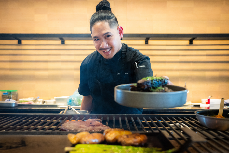 A smiling chef with hair pulled up in a bun leans over a large commercial grill, extending a round serving dish toward the camera. He wears a black chef's coat and apron, and several cuts of meat sizzle on the grill in the foreground. The kitchen has a warm wood-paneled backdrop.