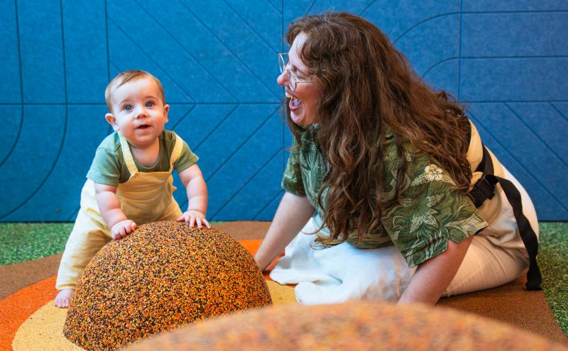 Infant leans against mound with parent smiling at them from the side.