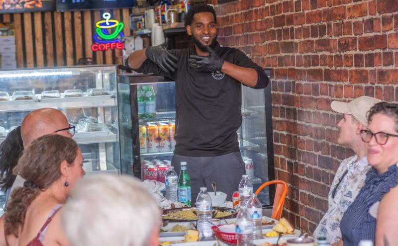 A guide at Culture Generation Food Tours in Kent speaks to a group of seated guests gathered around a table with plates of food inside a restaurant. The setting includes brick walls, a display case, and colorful menu signs.