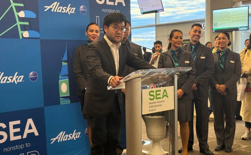 Port of Seattle Commissioner Sam Cho speaks at a podium in front of a banner celebrating Alaska Airlines' inaugural flight to London.