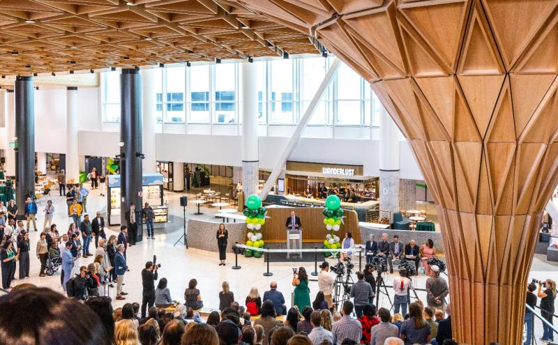 View from the Grand Stairs in the new C Concourse Expansion (CCE) building, looking down at the speaker during the CCE Grand Reveal Event. The Tree at C centerpiece of the new space is off to the right. People are seated on the amphitheater-style seating, listening to the speakers. 