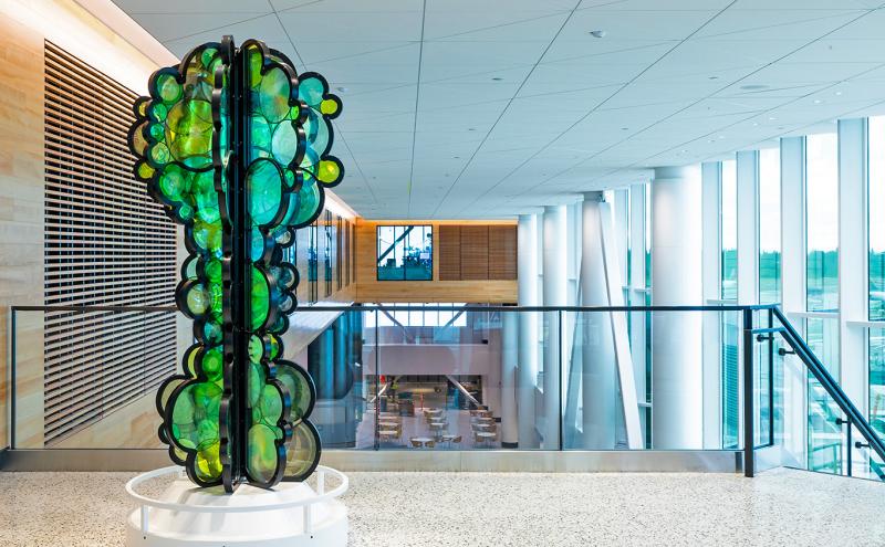 Artist Fumi Amano’s sculpture “Shared Breath” is shown on Level 3 of the C Concourse Expansion. The artwork consists of layered, circular green and yellow glass-like forms arranged vertically on a white base. It is displayed in a bright interior space with wood paneling, large windows, and views of seating and structural elements below.