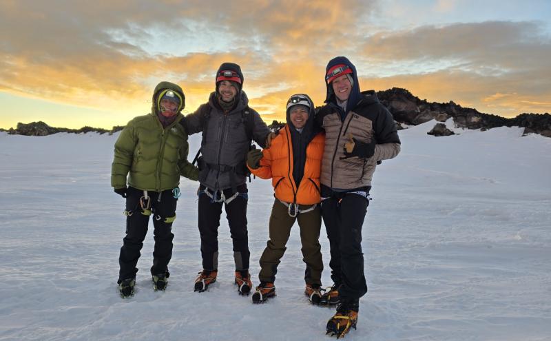 Four mountaineers in cold-weather climbing gear stand together on a snow-covered glacier at sunrise, smiling with a rocky volcanic ridge behind them.