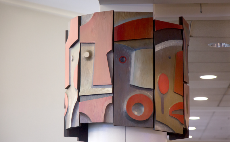 Two sides of the sculpture showing the wooden pieces projecting outward at different depths, giving the mask forms physical relief. 