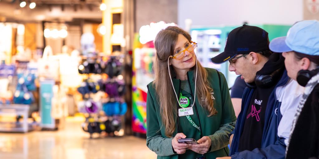 Volunteer in green with "ask me" button assists two travelers.