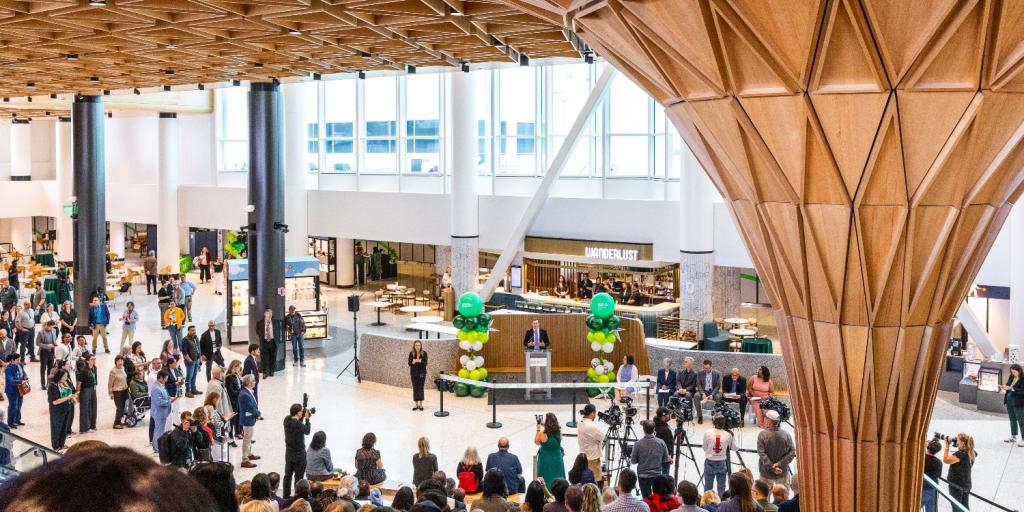 View from the Grand Stairs in the new C Concourse Expansion (CCE) building, looking down at the speaker during the CCE Grand Reveal Event. The Tree at C centerpiece of the new space is off to the right. People are seated on the amphitheater-style seating, listening to the speakers. 