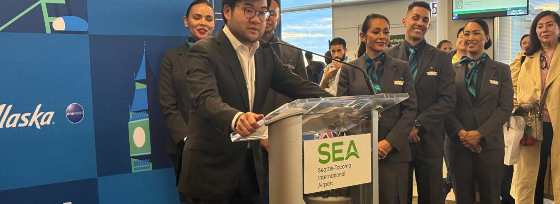 Port of Seattle Commissioner Sam Cho speaks at a podium in front of a banner celebrating Alaska Airlines' inaugural flight to London