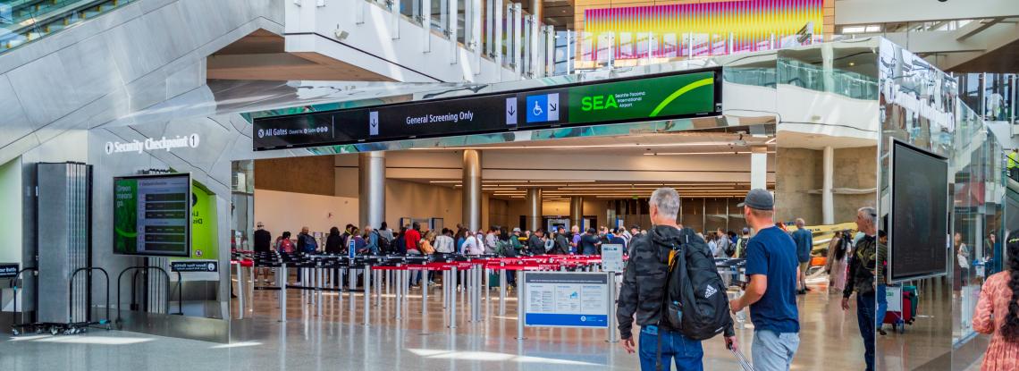 Two travelers walk towards Checkpoint 1, one with roller bag carry-on