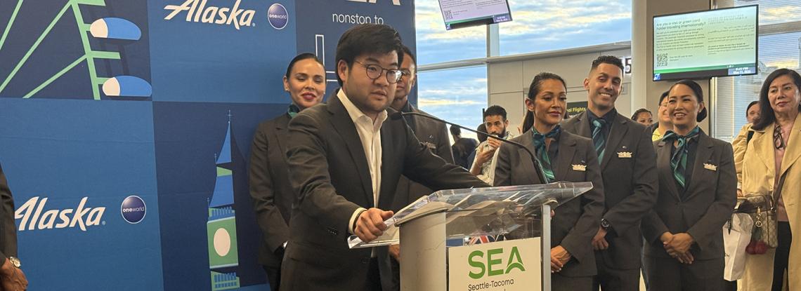 Port of Seattle Commissioner Sam Cho speaks at a podium in front of a banner celebrating Alaska Airlines' inaugural flight to London.