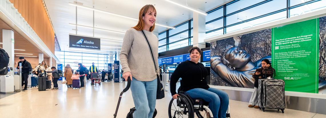 A traveler using a cane and traveler in a wheelchair walk through the terminal at SEA.
