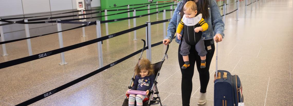 A mom walks in the terminal pushing a suitcase and a toddler in an umbrella stroller while also wearing a baby in a carrier.