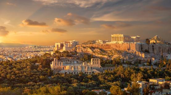 Panoramic view of the Acropolis of Athens, Greece, including the Parthenon and surrounding ancient ruins illuminated by warm evening sunlight, with the city of Athens stretching into the distance