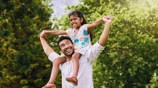 A girl rides on shoulders of her dad as they run through a park