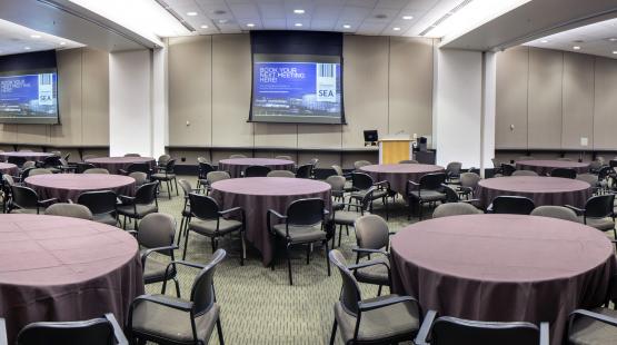 Panoramic view of the International Auditorium conference room. The room is set in 25 banquet rounds, with 7 chairs each. There are brown table cloths on each round table. All Airwalls are open, all three projectors are turned on, and all three dropdown projectors screens are displaying content.