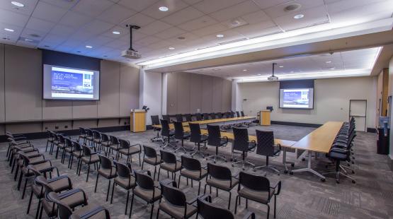 View from the London entry doorway looking into the International A conference room. The room is set in a u-shape, with rows of theater style seating behind it, facing the Amsterdam dropdown screen. Two projectors are turned and both projector screens are displaying content