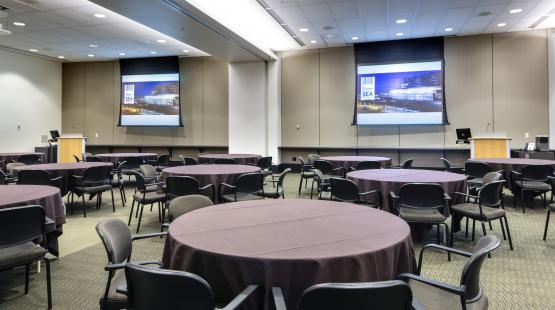View from the London entry doorway looking into the International B conference room. The room is set with Round tables with chairs, and each table is covered in a brown tablecloth. Two projectors are turned and both projector screens are displaying content.