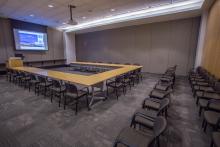 View from the entry doorway looking into the Beijing Conference room that is in a u-shape room set and two rows of theater style seating behind it. The projector is turned and the projector screen is displaying content
