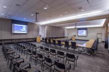 View from the London entry doorway looking into the International A conference room. The room is set in a u-shape, with rows of theater style seating behind it, facing the Amsterdam dropdown screen. Two projectors are turned and both projector screens are displaying content
