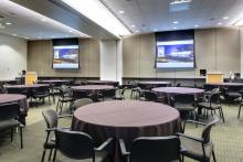 View from the London entry doorway looking into the International B conference room. The room is set with Round tables with chairs, and each table is covered in a brown tablecloth. Two projectors are turned and both projector screens are displaying content.