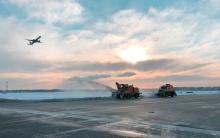 Snowplows at SEA plow the runways while a plane takes off with a sunset.