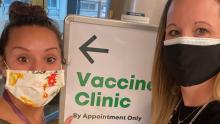 Two femal masked staff members along side sign pointing to a COVID-19 Vaccine Clinic at SEA Airport April 2021