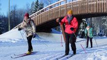 Photo of three cross country skiers on snow-covered trail cross under a pedestrian arched bridge with sunny blue skiesskies
