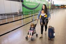 A mom walks in the terminal pushing a suitcase and a toddler in an umbrella stroller while also wearing a baby in a carrier. 