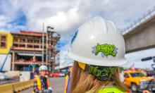 Worker with a construction hat that has the Upgrade SEA logo while a construction project is in progress in the background. 